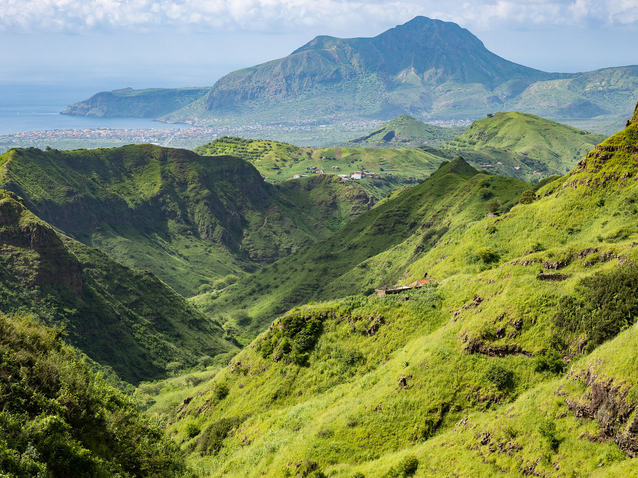 Paisagem com montanhas verdejantes que se estendem até ao horizonte, onde se pode ver a cidade e o mar azul ao longe