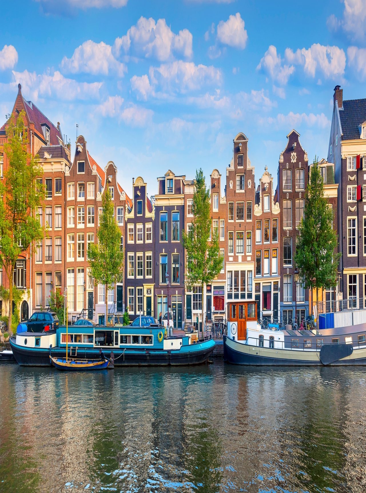 Canal de Amsterdão com pessoas a caminhar pela margem, barcos, e edifícios típicos, com um céu azul 