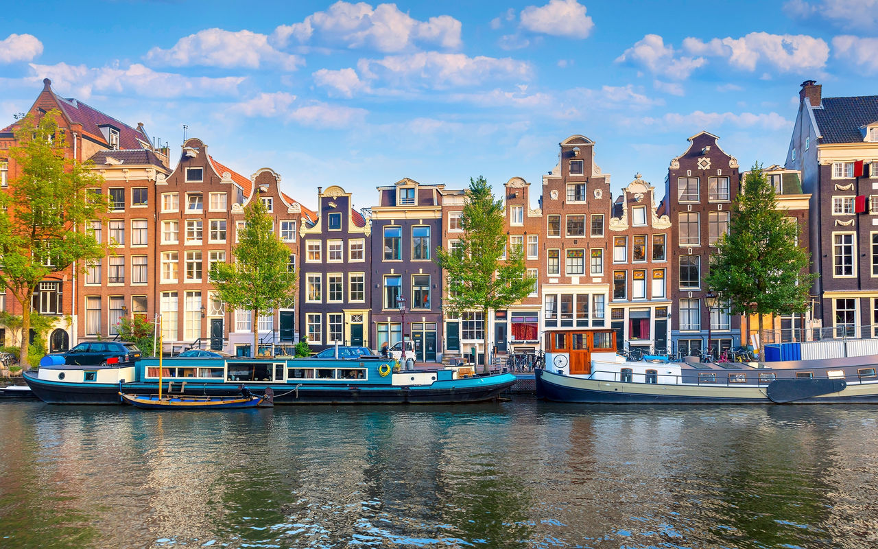 Canal de Amsterdão com pessoas a caminhar pela margem, barcos, e edifícios típicos, com um céu azul 