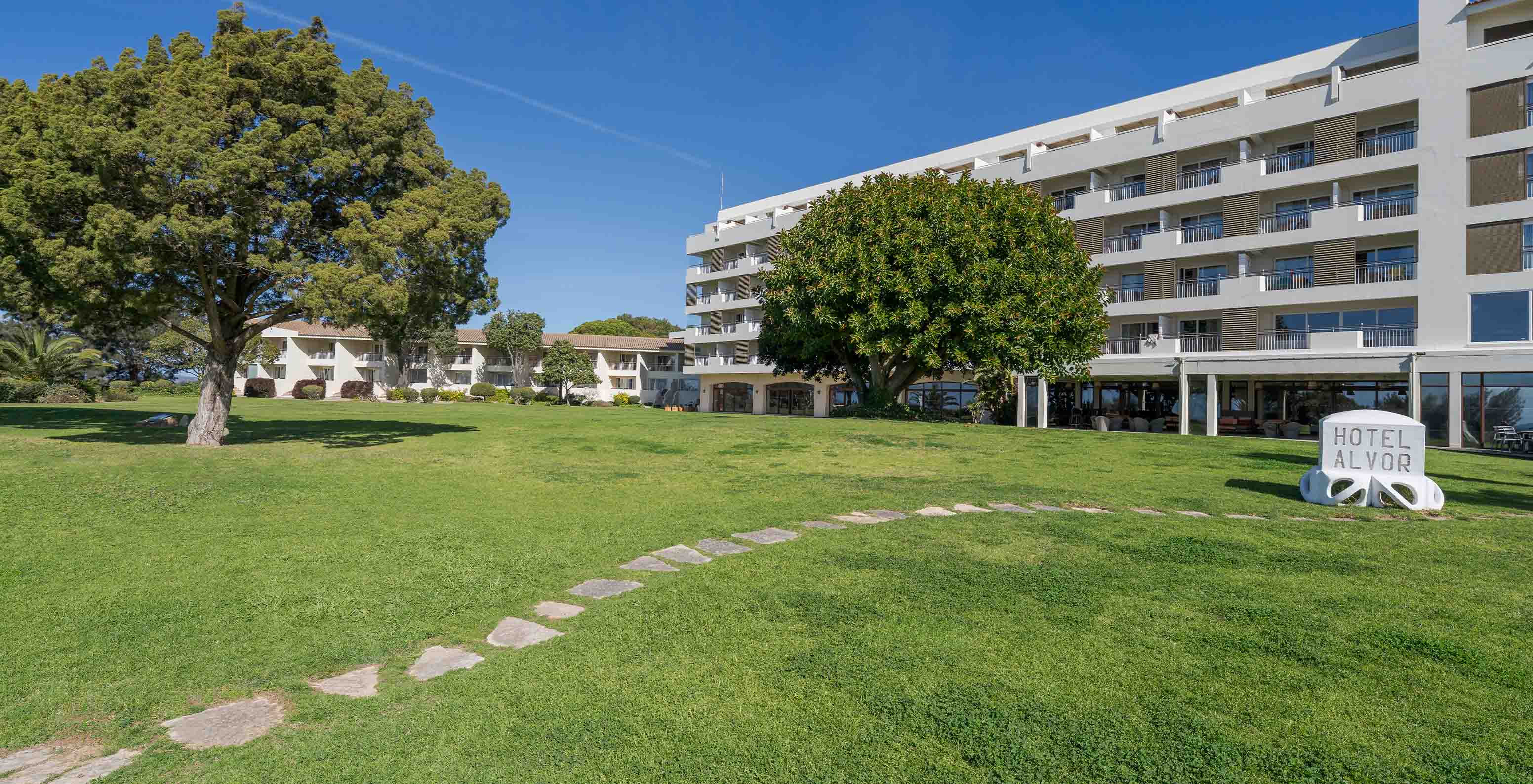 Garten mit Bäumen des Pestana Alvor Praia, mit Hotelgebäuden im Hintergrund und einem Steinweg