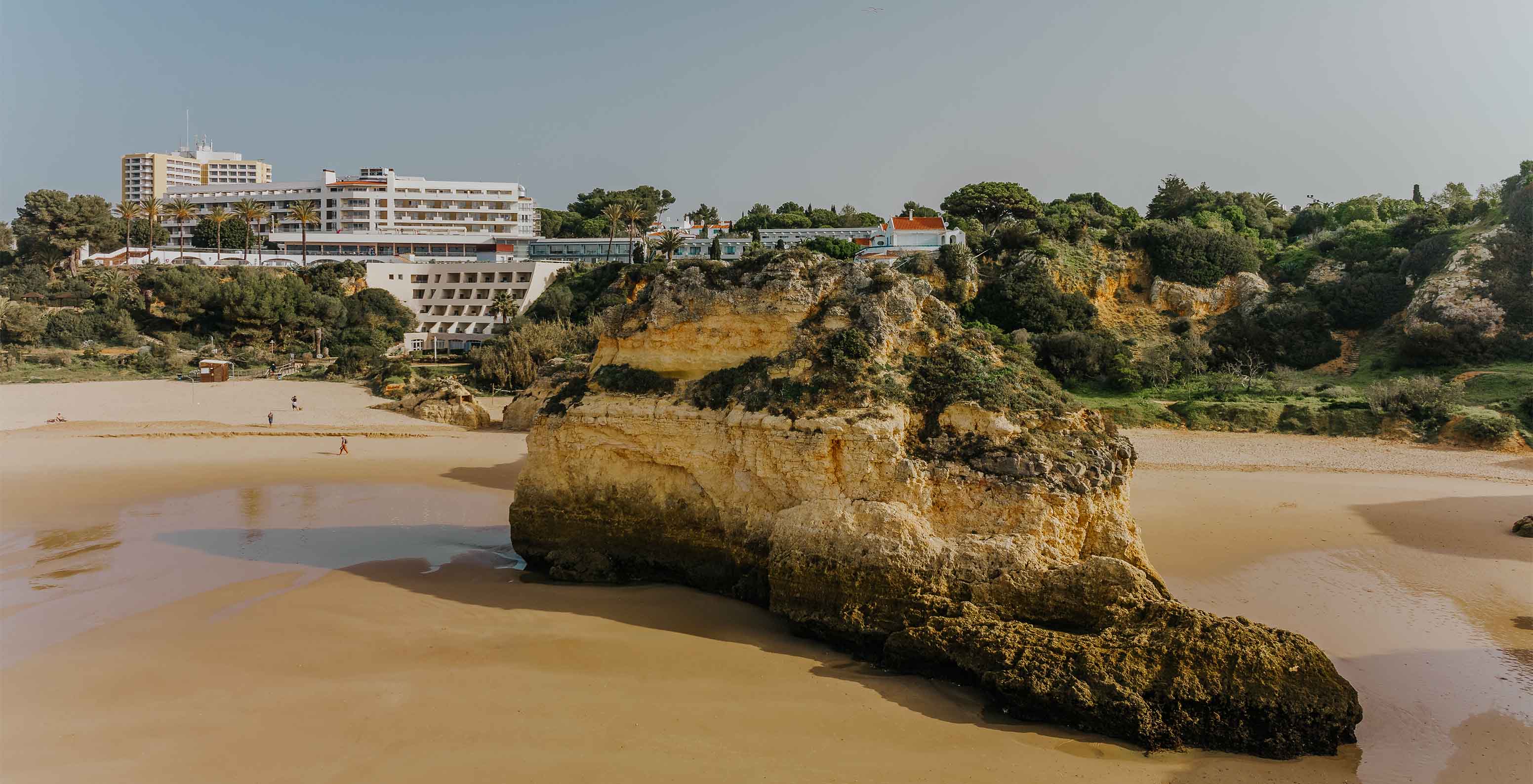 Strand von Alvor mit Felsen, Sand, Vegetation, der Klippe und Gebäuden im Hintergrund, mit klarem Himmel