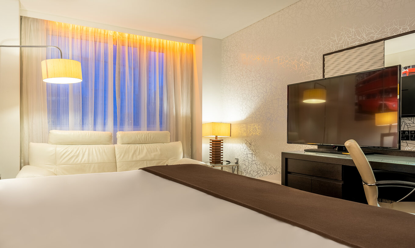Das Deluxe King im Pestana Chelsea Bridge hat Bett, Schreibtisch mit TV und Stuhl, Sofa und Fenster zur Stadt