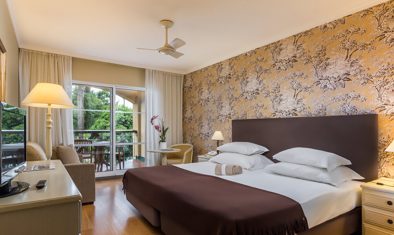 The Studio Classic at Pestana Village has a double bed with headboard and brown bedspread, floral wallpaper and balcony
