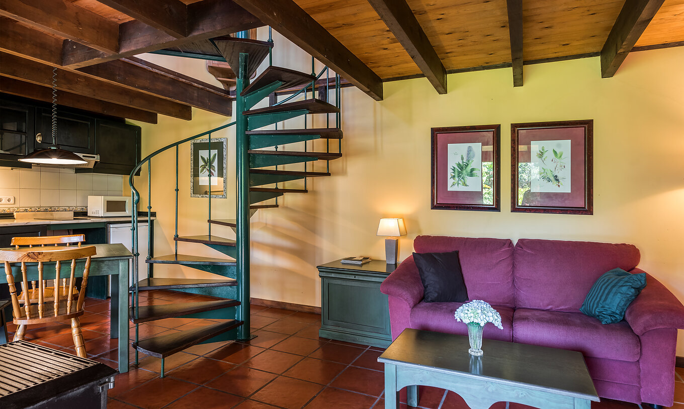 The Duplex Villas of Pestana Quinta do Arco have a sofa with a side table, kitchen, dining table, and metal stairs