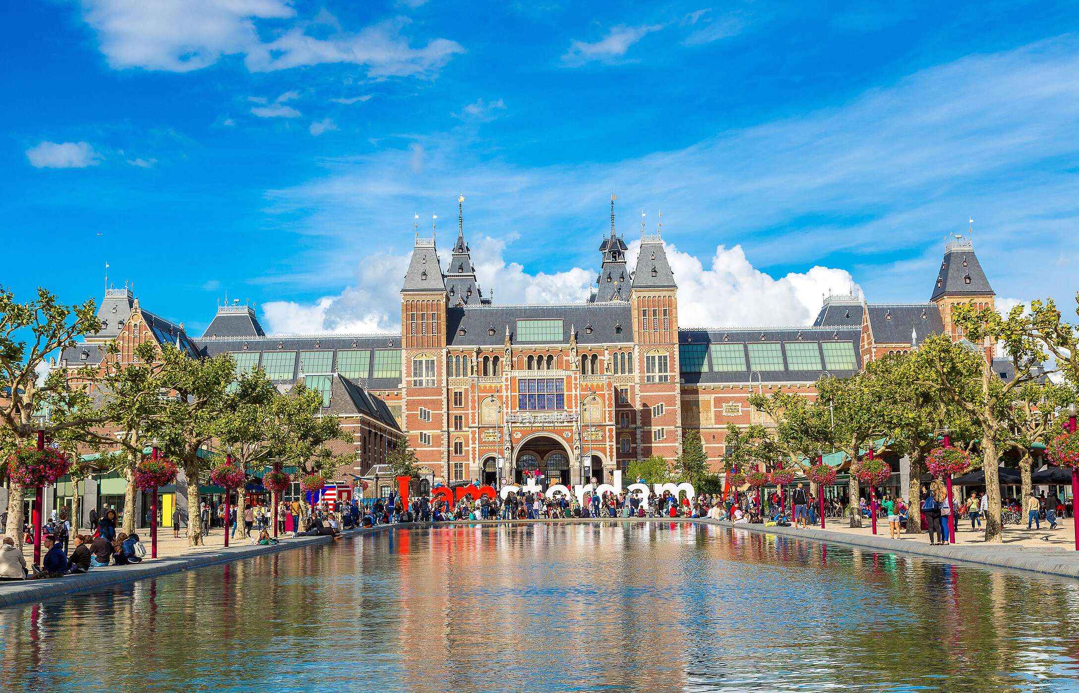 Facade of the famous Rijksmuseum in Amsterdam, Netherlands, with people walking, trees, and a lake in the foreground.