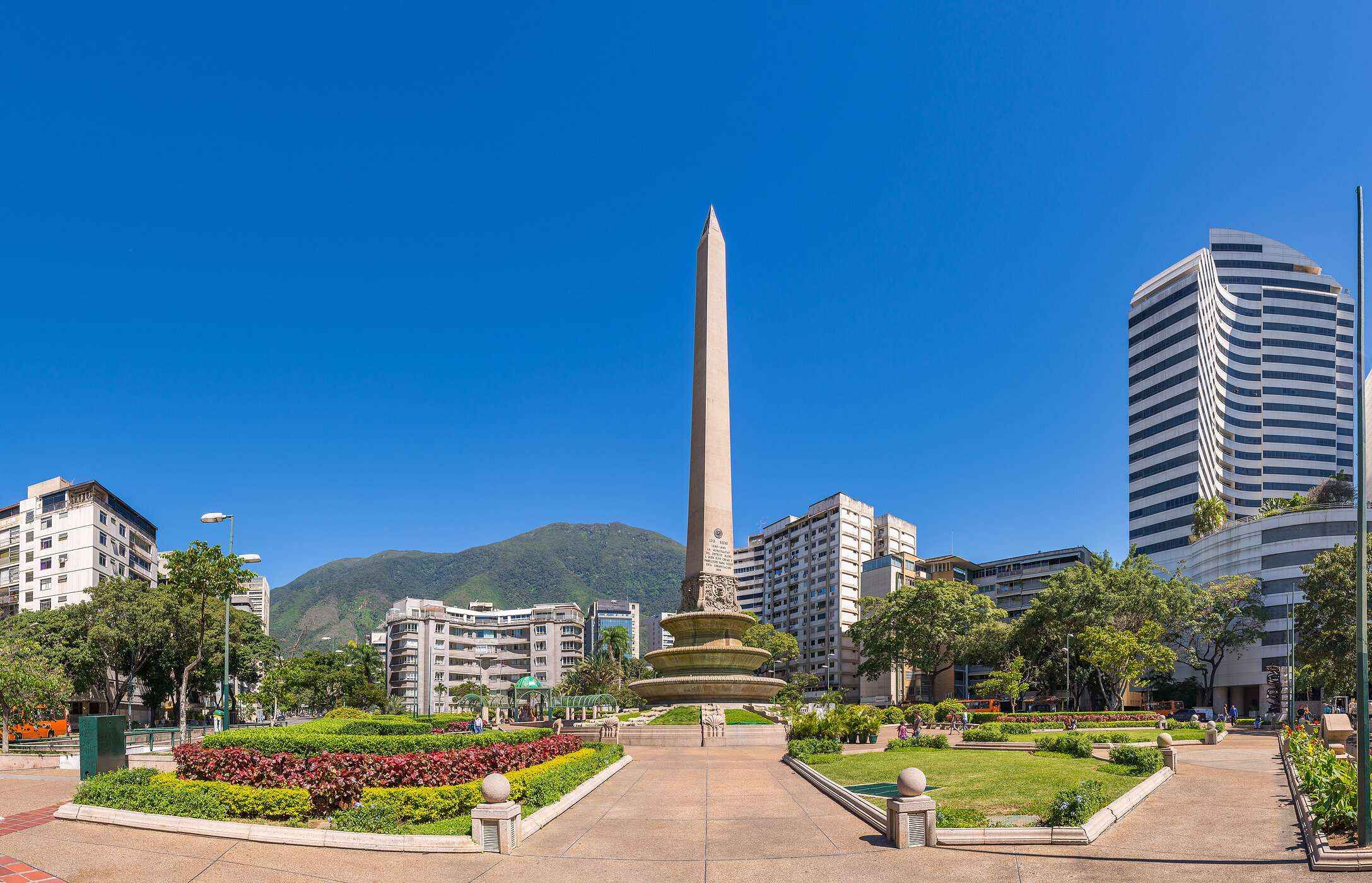 Monumento Obelisco, una gran columna de piedra en medio de un parque con flores, rodeada de edificios altos en Venezuela.