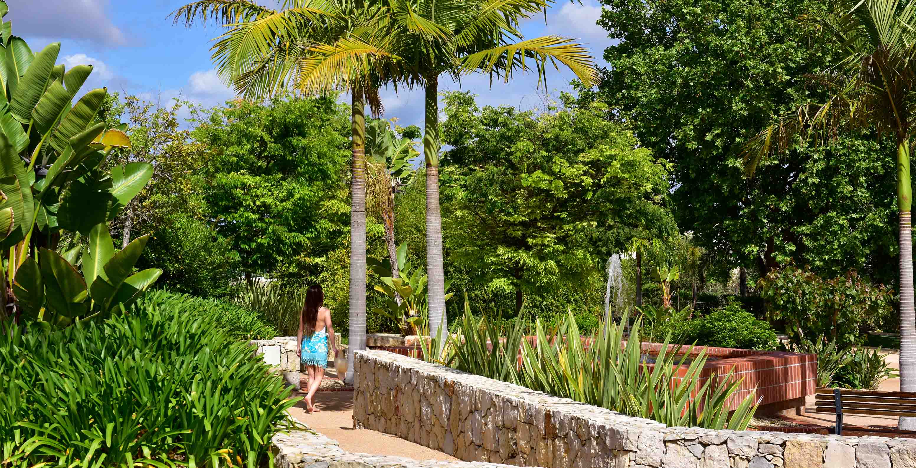 Jardin avec de l'herbe, des palmiers et divers arbres, avec une dame sur le chemin bordé de murs en pierre