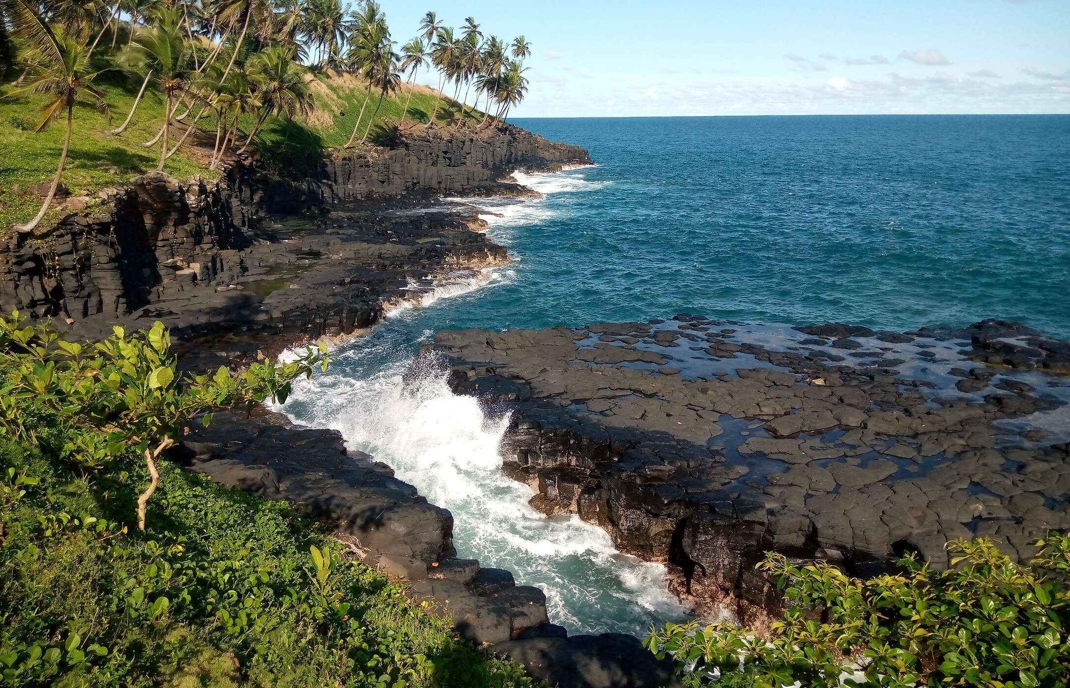 Vue sur la Bouche de l'Enfer à São Tomé et Príncipe, avec une mer agitée, des rochers noirs et de la végétation autour.