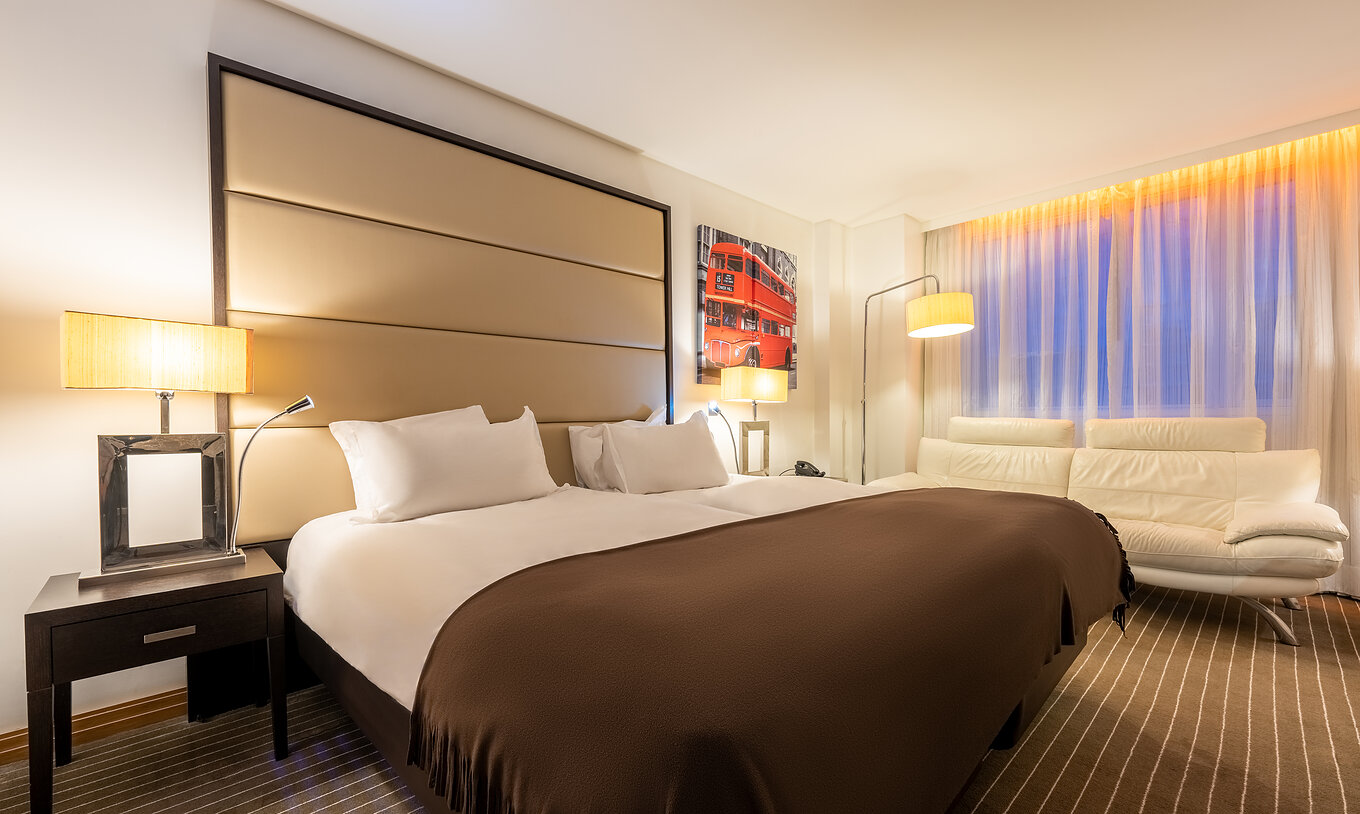 Le Premium Twin du Pestana Chelsea Bridge a deux lits, table de nuit, canapé, lampes et fenêtres avec rideaux