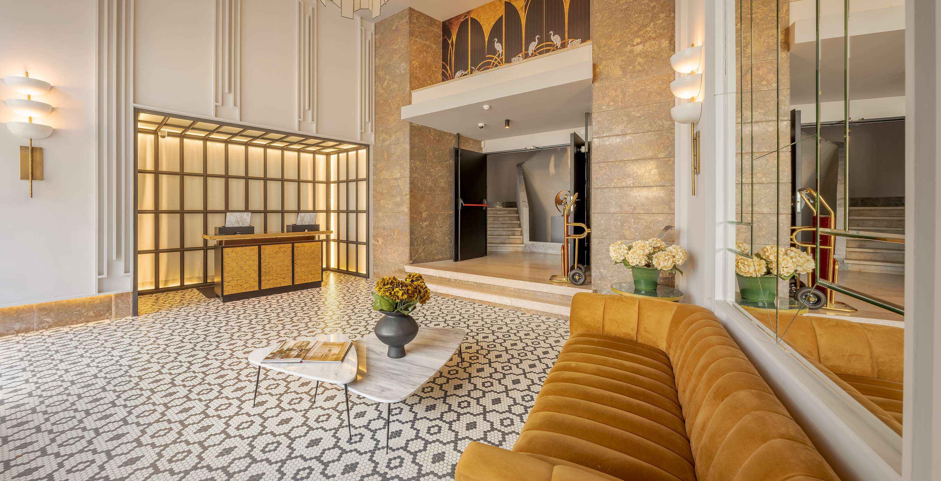 Lounge da receção com um casal e uma criança a andar, espelhos, um sofá, mesas de apoio com vasos de flores e receção