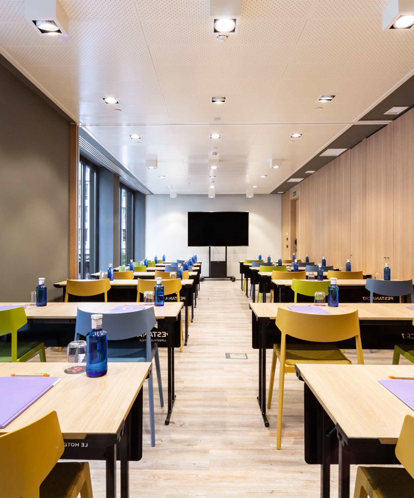 Sala de eventos e reuniões com mesas de madeira com cadeiras amarelas, verdes e azuis, com televisão e paredes de madeira