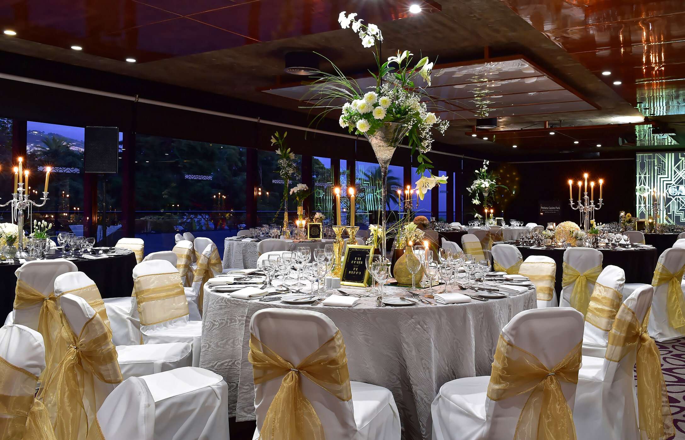 Casamento em sala com decoração moderna, com mesas redondas com arranjos florais altos e iluminação suave e elegante