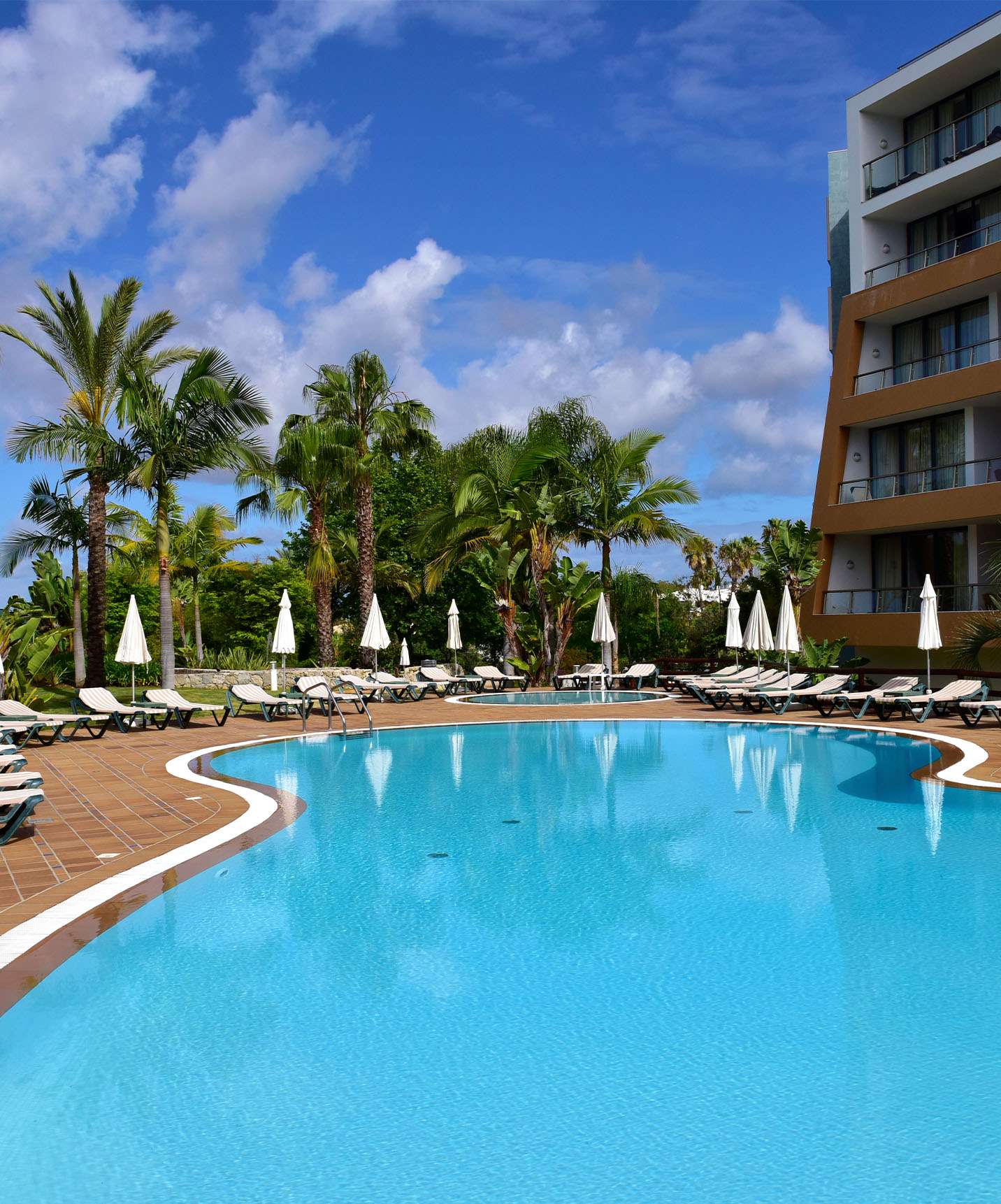 Piscinas exteriores com espreguiçadeiras, chapéus de sol e palmeiras, com o edifício do hotel na lateral