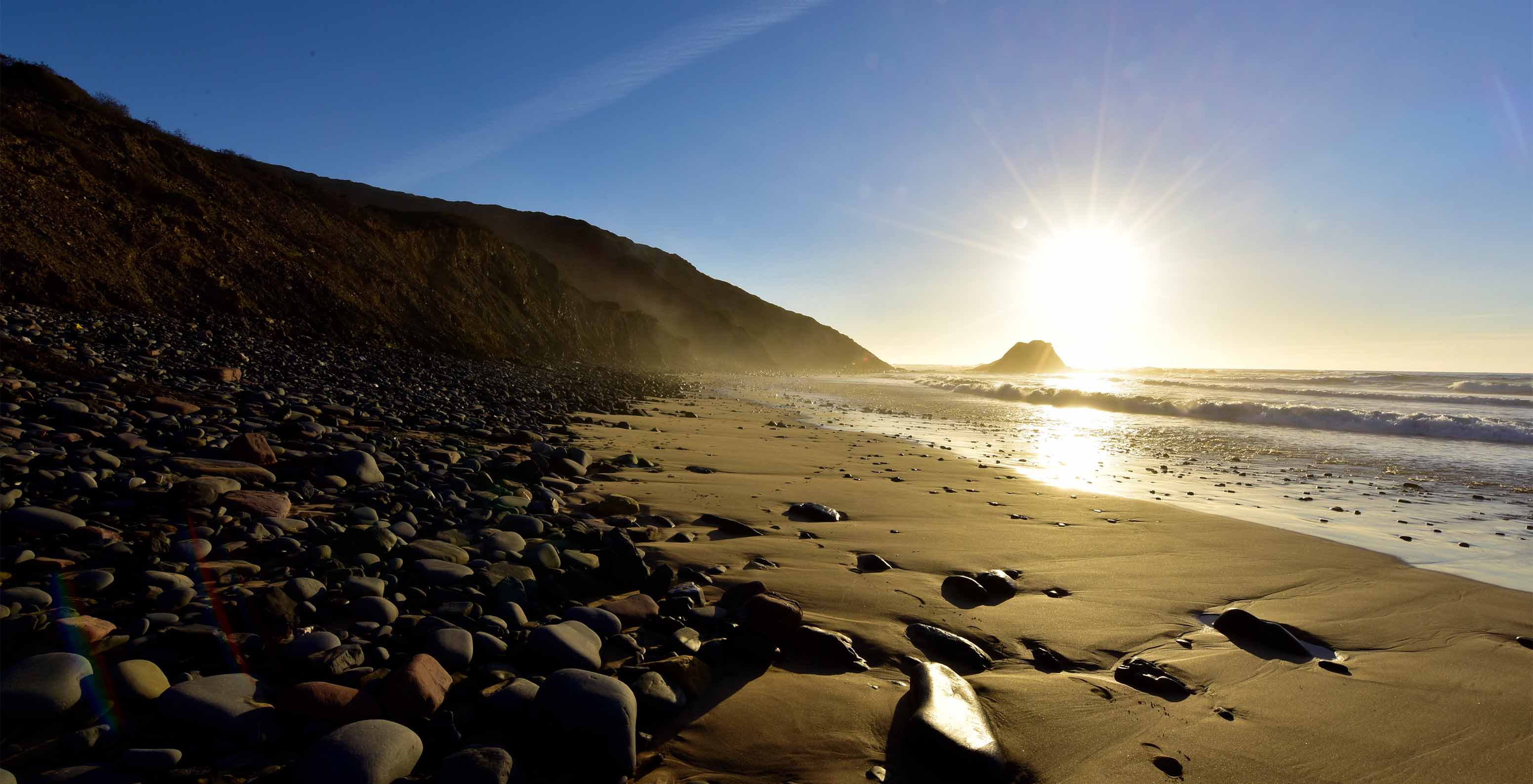 Praia no nascer do sol, com a luz a refletir no mar, pedras na areia e uma falésia atrás