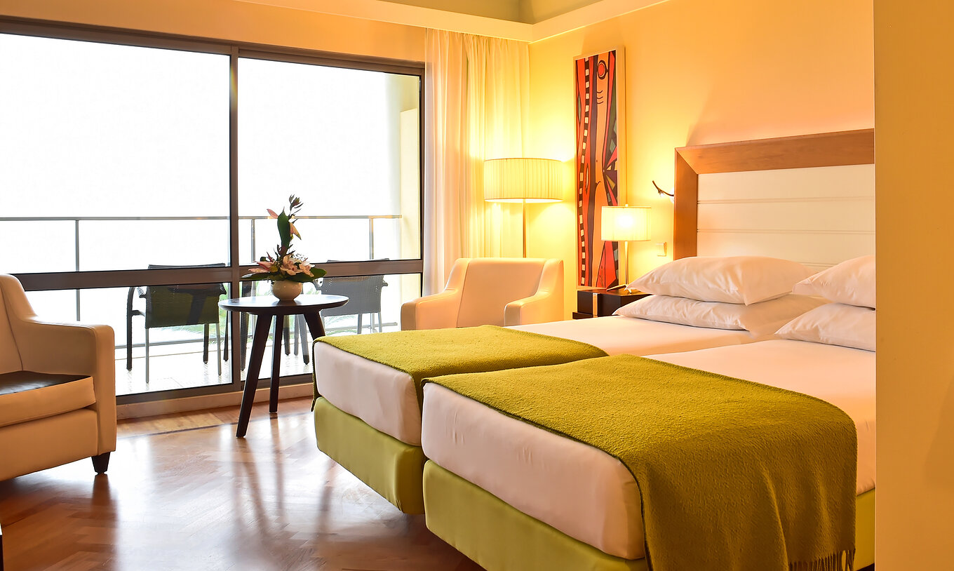 O Superior do Pestana Promenade tem duas camas com uma colcha verde e uma janela grande para a varanda