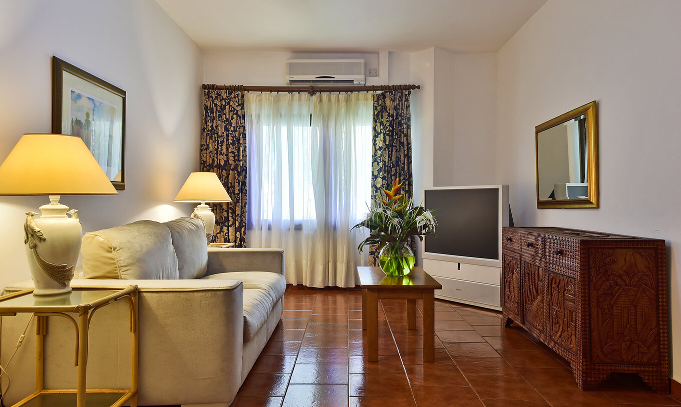 A Suite do Pestana Miramar São Tomé tem uma sala de estar com sofá, televisão e uma mesa com uma jarra de flores