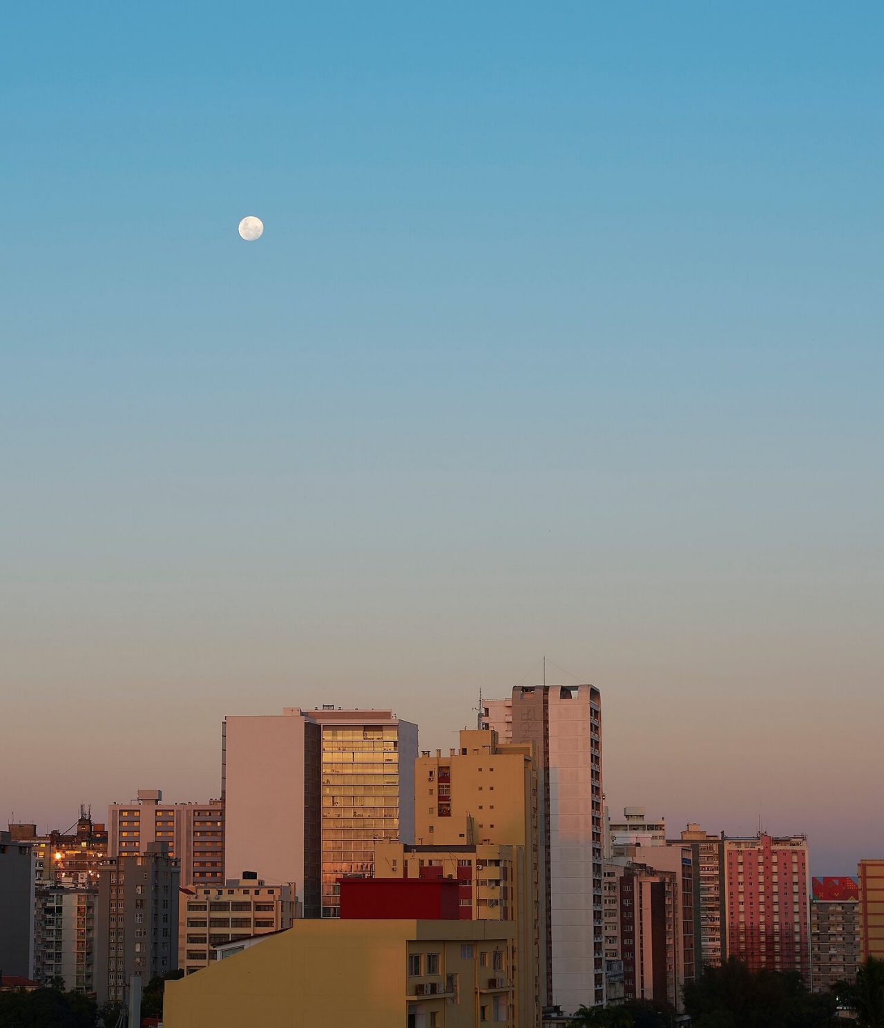 Blick von oben auf mehrere Gebäude in Maputo, mit Dämmerlicht auf ihnen und dem Mond hoch am Himmel