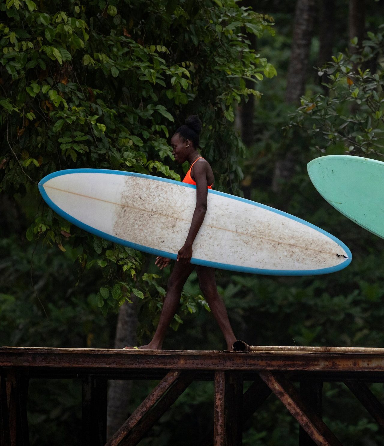 Frau in São Tomé, die über eine Eisenbrücke geht, eine Surfbretter in der Hand hält, mit Vegetation dahinter