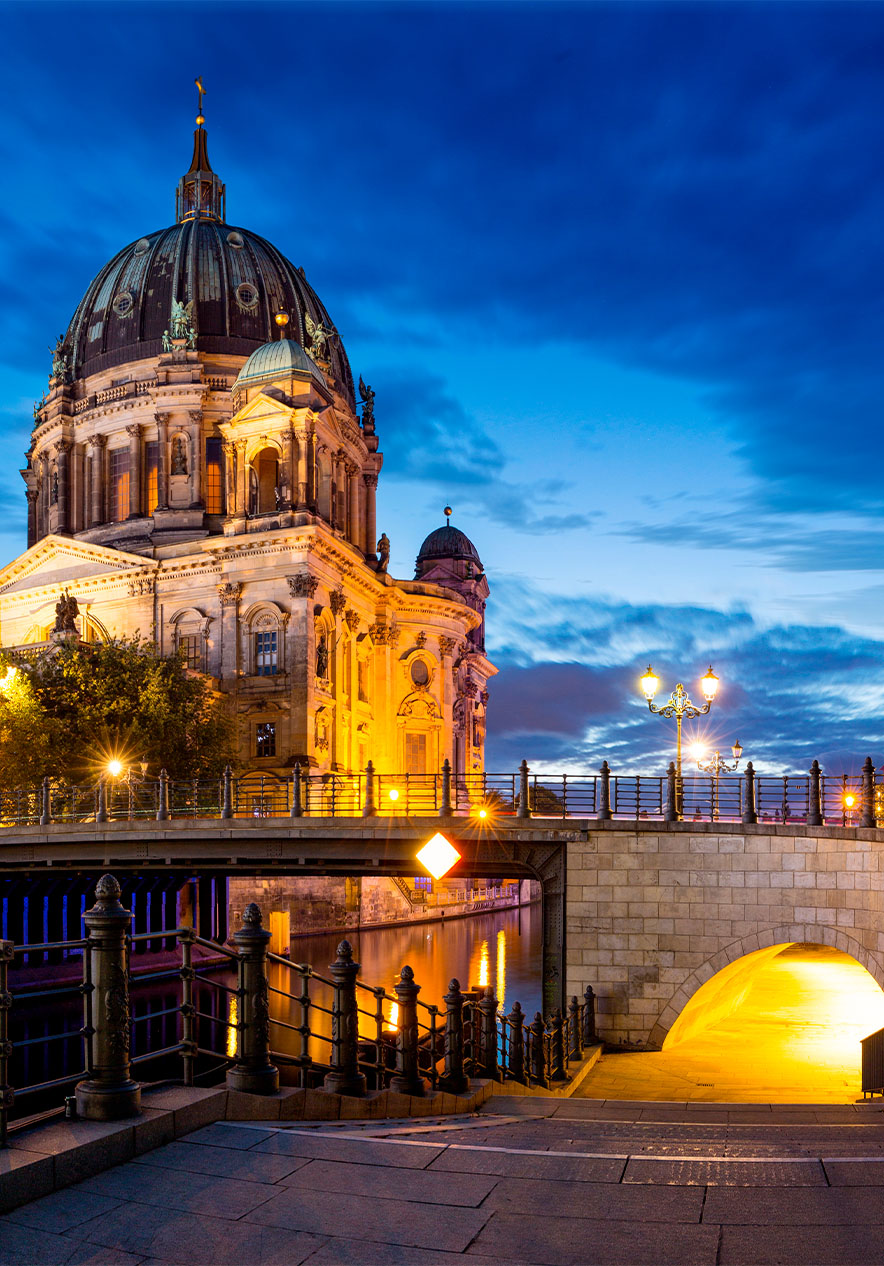 Berliner Dom bei Dämmerung beleuchtet, mit einer Brücke und modernen Gebäuden, die sich im Fluss spiegeln