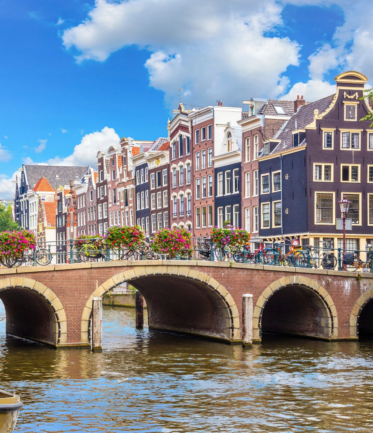 Fußgängerbrücke über einen Kanal in Amsterdam, mit geparkten Fahrrädern, bunten Blumen und Gebäuden im Hintergrund