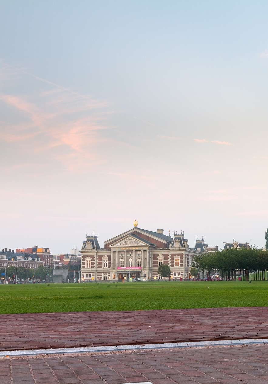 Blick auf das Gras und den gepflasterten Bereich des Museumplein in Amsterdam, mit Bäumen und dem Museum im Hintergrund