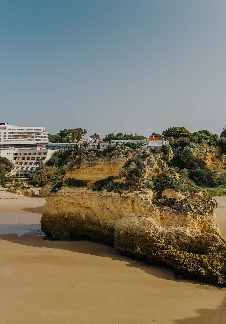 Strand von Alvor mit Felsen, Sand, Vegetation, Klippe und Gebäuden im Hintergrund unter klarem Himmel
