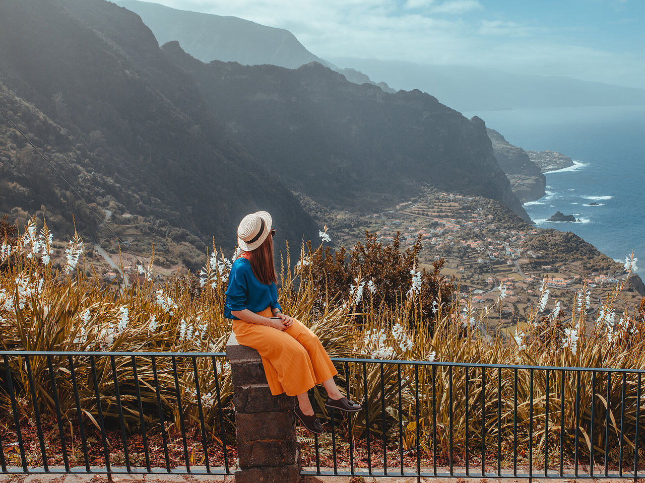 Frau sitzt auf einer Mauer und genießt den Blick auf eine Fajã auf der Insel Madeira, mit dem Meer drumherum