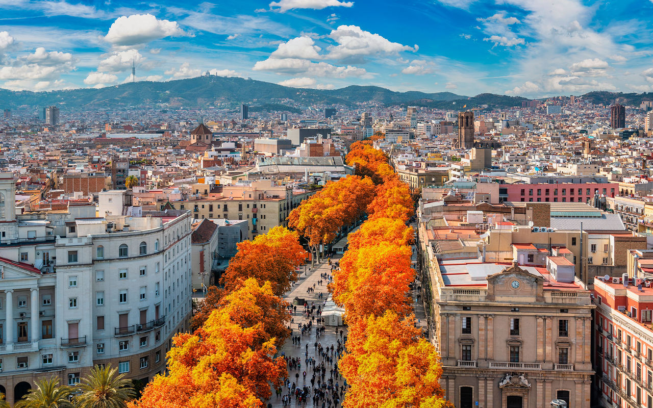 Vista aérea sobre la ciudad de Barcelona, con personas paseando, árboles coloridos con hojas de otoño y varios edificios