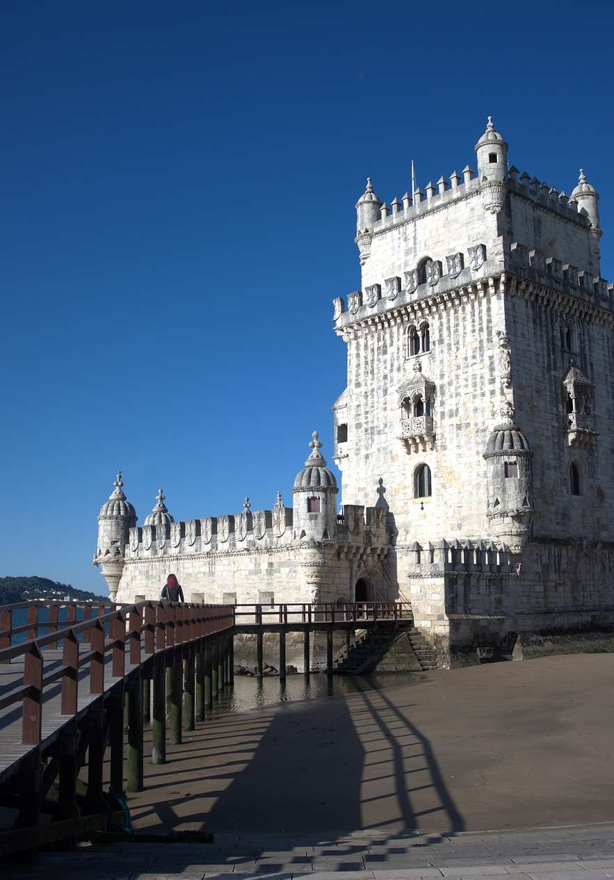 Torre de Belém con pasadizo de madera al lado, arena oscura, el río Tajo detrás de la torre y árboles alrededor