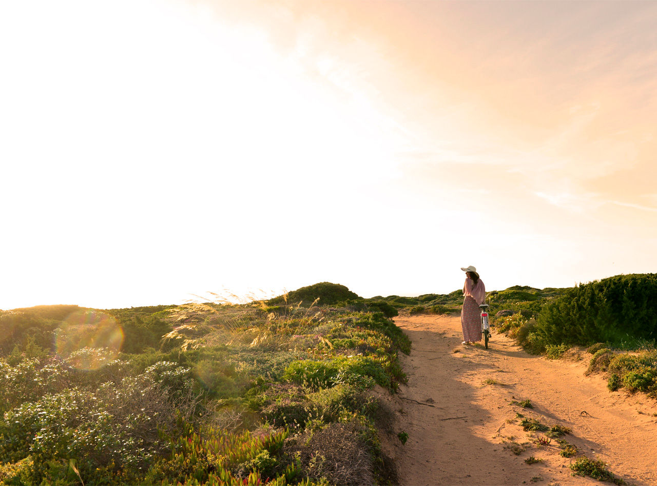 Femme faisant du vélo sur un chemin de terre à travers les dunes, avec un vélo des Pousadas de Portugal