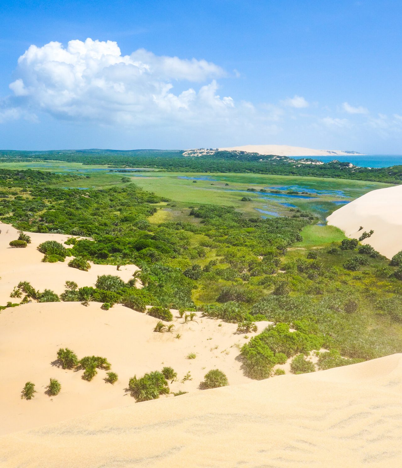 Vista aérea de uma paisagem desértica com dunas de areia branca, uma lagoa e águas cristalinas do oceano no fundo