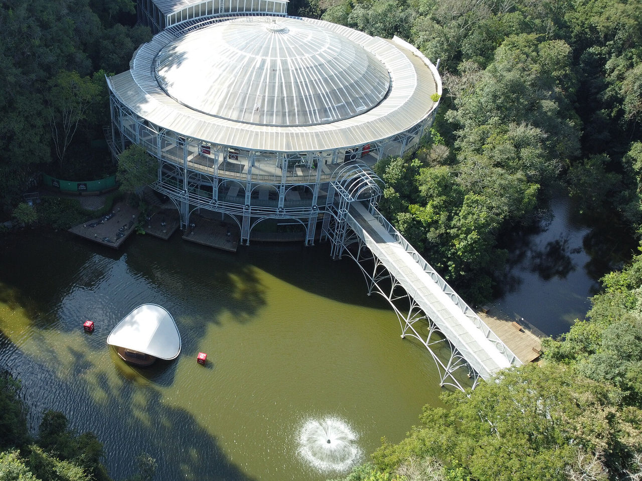 Vista aérea da ópera de Arame, com o a estrutura redonda icônica, envolta na natureza, com uma ponte e um lago