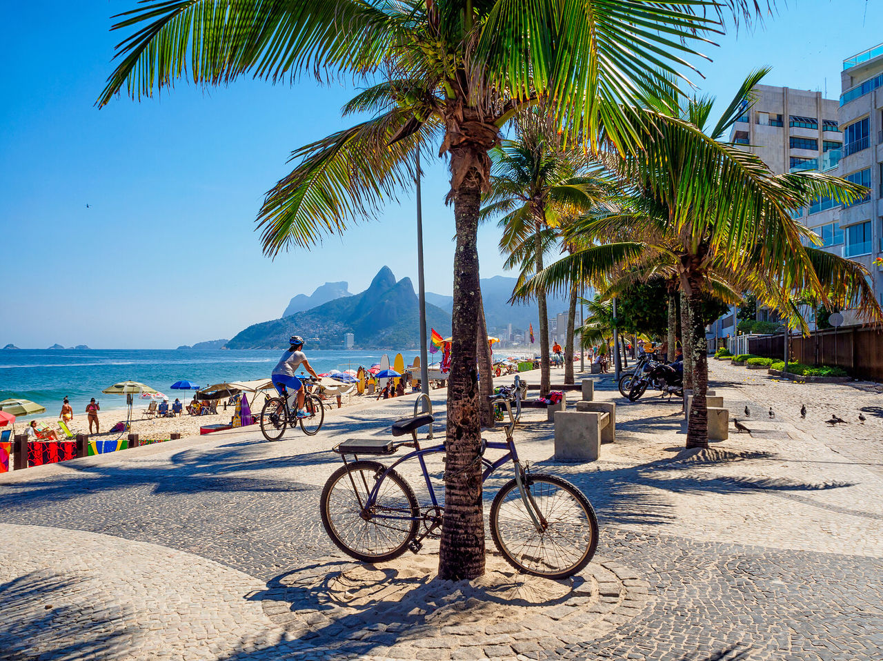 Vista da praia de Ipanema, com pessoas a apanhar sol, ciclistas, e o morro dos Dois Irmãos em segundo plano