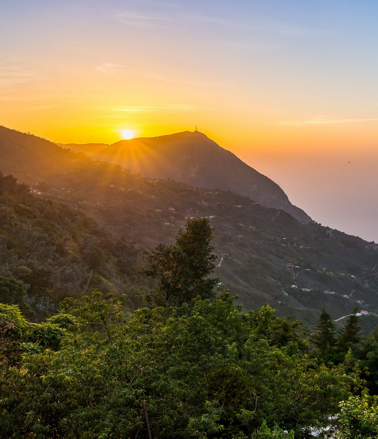 Vista aérea de uma montanha em Caracas, com vegetação e algumas habitações, com um céu colorido no fundo