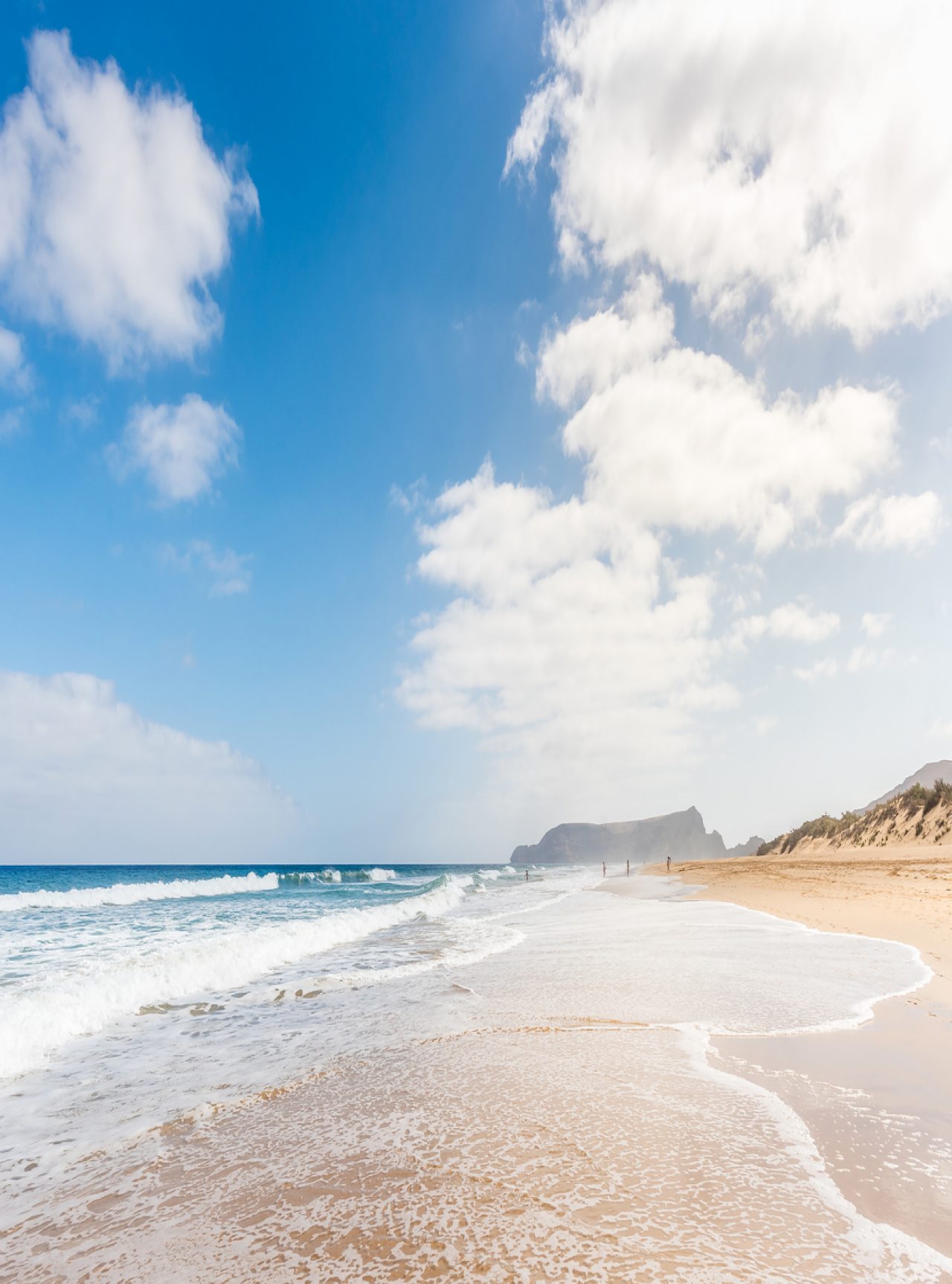 Praia extensa em Porto Santo com areia clara, ondas calmas, e um céu azul com alguma nuvens