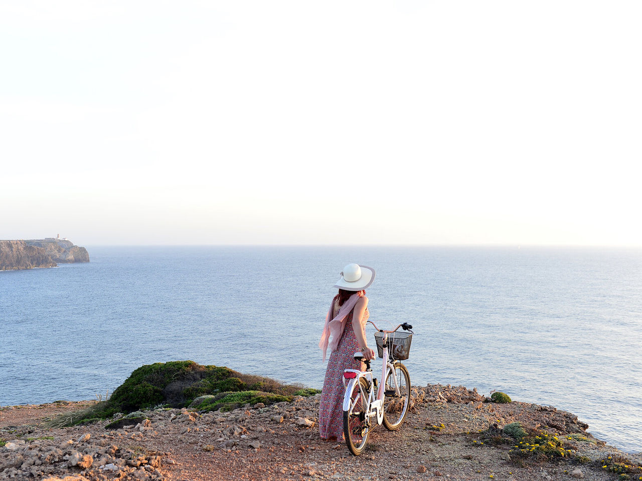Mulher jovem de chapéu e bicicleta na mão, enquanto aprecia a vista para o mar numa falésia no Algarve
