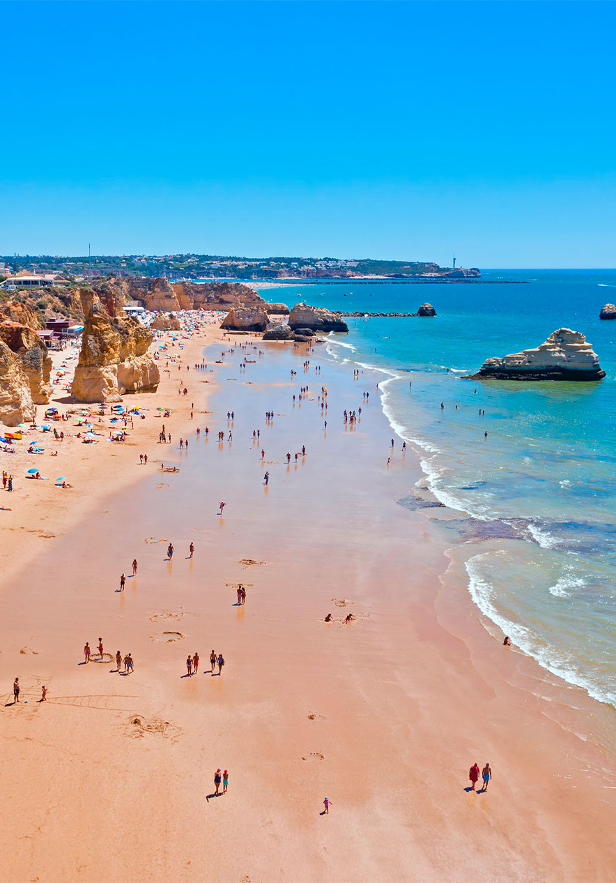 Vista aérea da Praia da Rocha com guarda-sóis, falésias, pessoas, rochas e o mar, com o céu limpo