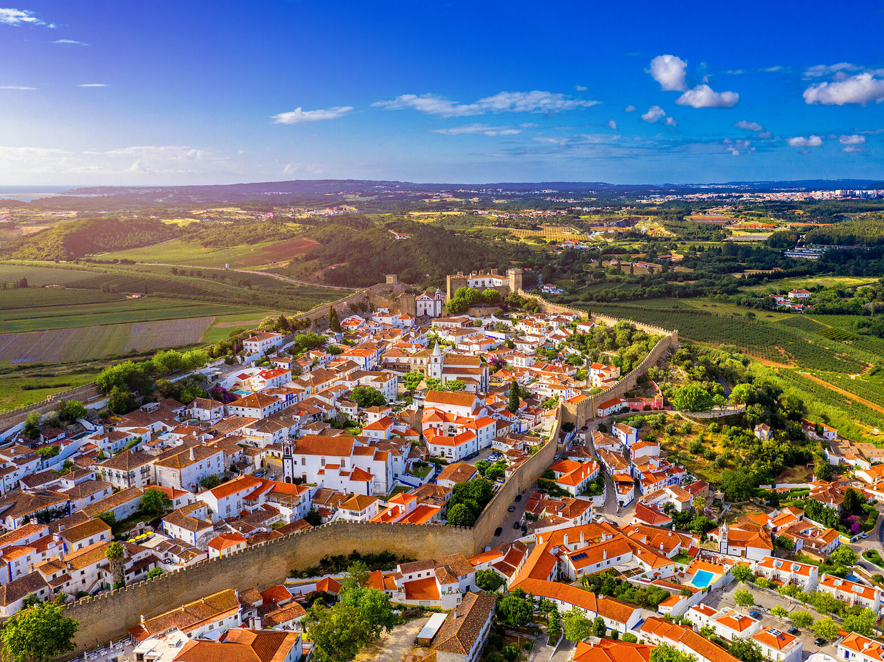 Vista aérea de uma vila rodeada por campos verdes e com parte da vila cercada por muralhas