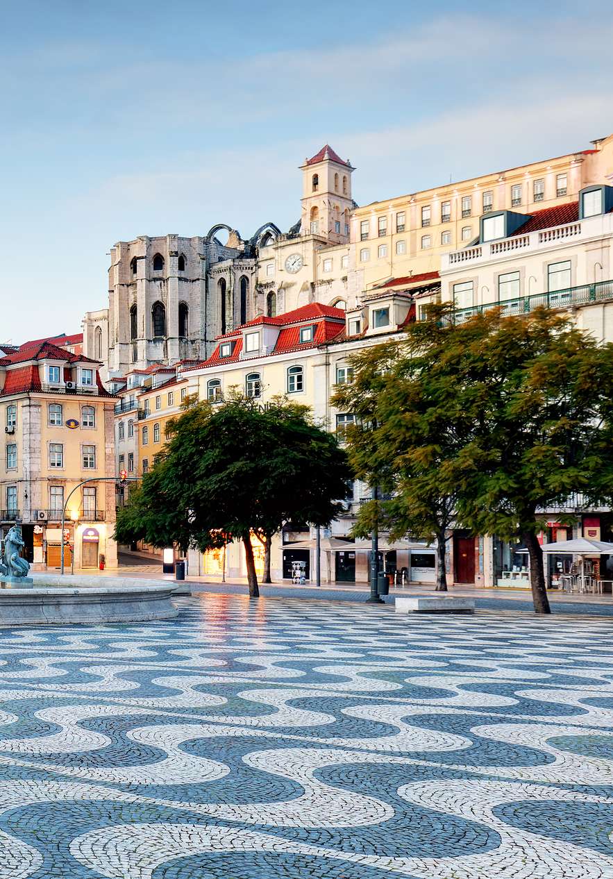 Vista para a Praça do Rossio com calçada portuguesa, uma fonte central com estátuas e vários prédios ao redor