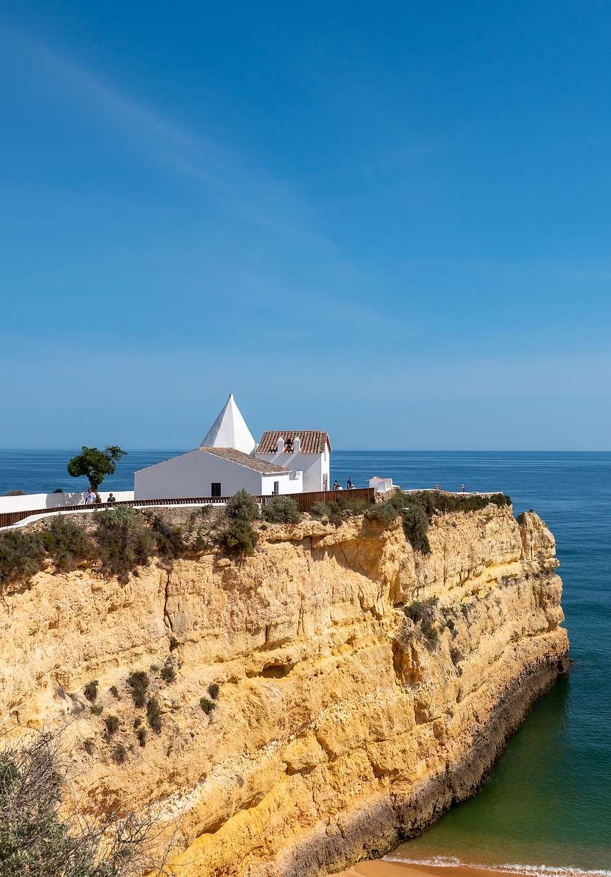View over the cliff of Senhora da Rocha beach with a small white house on top of the cliff and a calm sea in the background