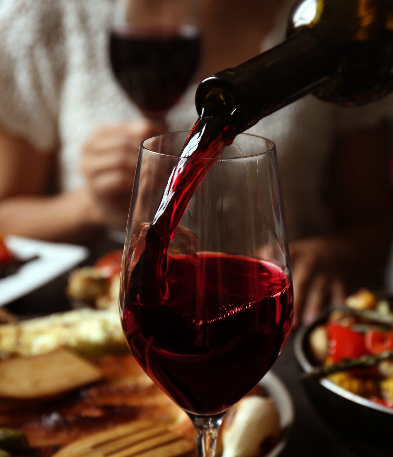Port red wine being served in a tall glass, with a person behind drinking wine and dishes of food